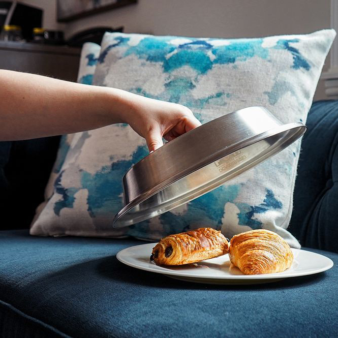 a plate with two croissants placed on a sofa and a hand lifting the cover.